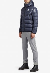 Uomo che indossa un giubbotto imbottito con cappuccio blu navy, pantaloni grigi chiari con polsini e stivali neri stringati, mentre è in piedi su uno sfondo bianco uniforme.