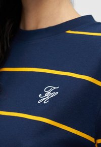 Μπλε ναυτικό βαμβακερό μπλουζάκι με κίτρινες οριζόντιες ρίγες, στρογγυλή λαιμόκοψη και λευκό κεντημένο λογότυπο στην αριστερή πλευρά. Μαλακή υφή.