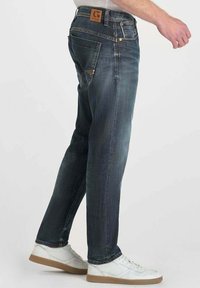 Jeans de mezclilla en un tono oscuro con un corte recto, que presentan un parche de cuero marrón, bolsillos laterales y costuras en contraste. Llevados con zapatillas blancas.