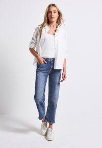 Femme avec de longs cheveux blonds portant une chemise blanche, un débardeur blanc, un jean bleu et des baskets blanches se tenant devant un fond blanc uni.