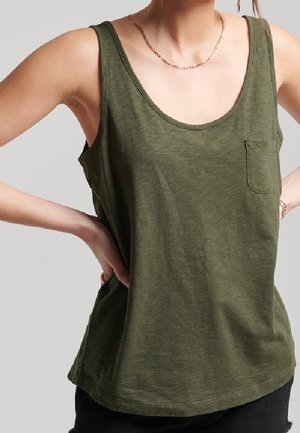 Kobieta w oliwkowo-zielonym, bezrękawnym topie z kieszenią z przodu, stojąca z rękami na biodrach na jednolitym tle.