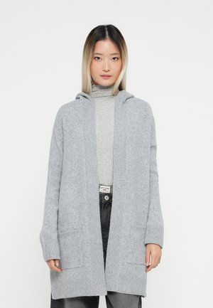 Kvinna med rakt, axellångt hår, iklädd en ljusgrå huva cardigan över en grå polotröja och mörkgrå jeans.