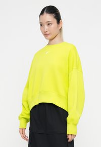 Svetlo neonsko rumeno oversized sweatshirt z zaobljenim ovratnikom, spuščeni rameni in majhnim belim logotipom Nike na prsih.
