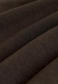 Brūns tekstils ar teksturētu austu struktūru, parādot locījumus un rievas, uzsverot tā auduma sastāvu un bagāto, tumšo krāsu.