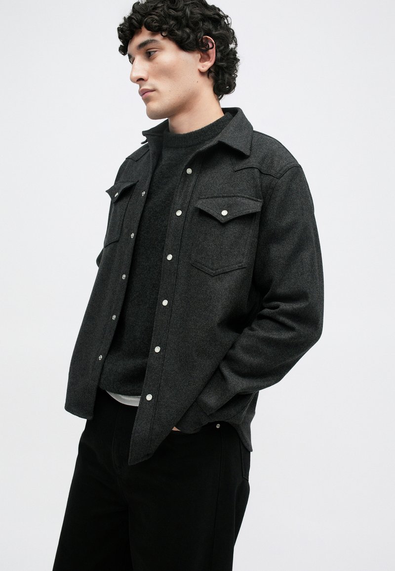 Gray wool shirt jacket with a structured design, snap buttons, two chest pockets, and a black sweater beneath, paired with black pants.