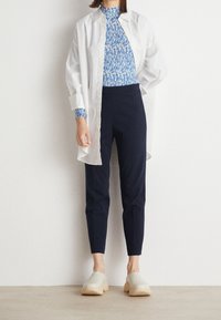 Kvinna iklädd en vit oversized knappskjorta, ett blåmönstrat linne, marinblå tapered byxor och ljusa slipskor, stående mot en vit vägg.