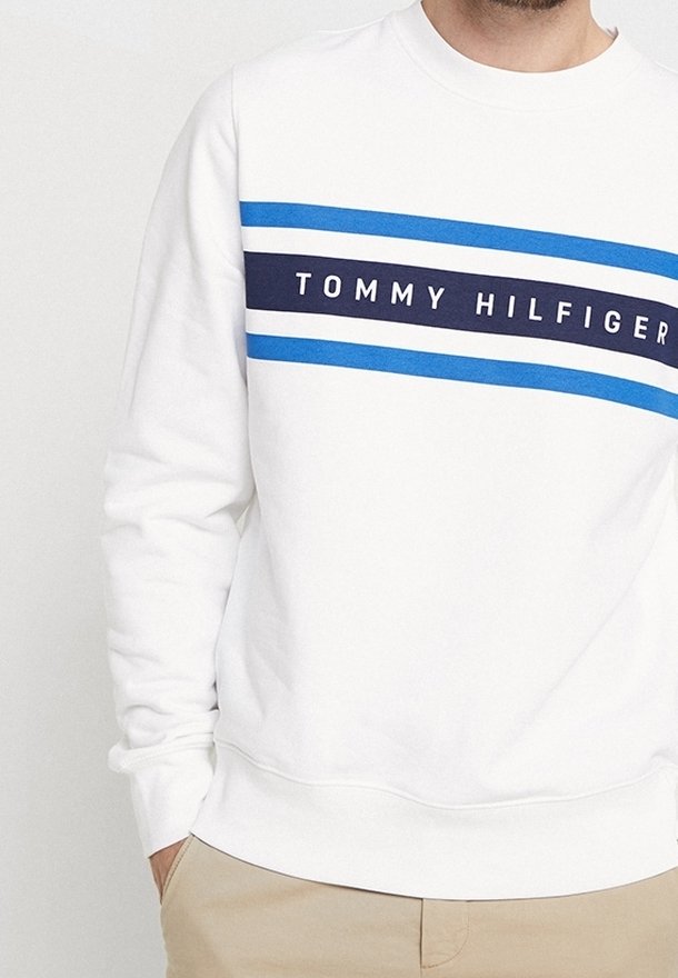 Sweat-shirt blanc avec des rayures bleues et le texte "Tommy Hilfiger" porté par une personne avec un pantalon beige, main dans la poche, visible du cou à la taille.