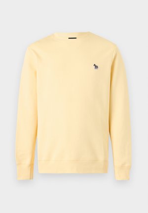 Mann trägt hellgelben Pullover mit kleinem Logo, dunkelblaue Jogginghose, weiße Sneaker, steht mit den Händen in den Taschen vor hellem grauem Hintergrund.