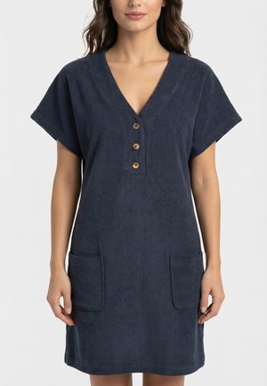 Mujer con vestido azul marino de manga corta con escote en V, tres botones de madera y dos bolsillos delanteros de parche, de pie contra un fondo liso.