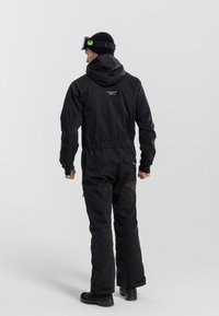 Czarny kombinezon narciarski z kapturem, regulowanymi mankietami i zapinanymi kieszeniami. Posiada logo z tyłu i jest wykonany z materiału odpornego na warunki atmosferyczne.