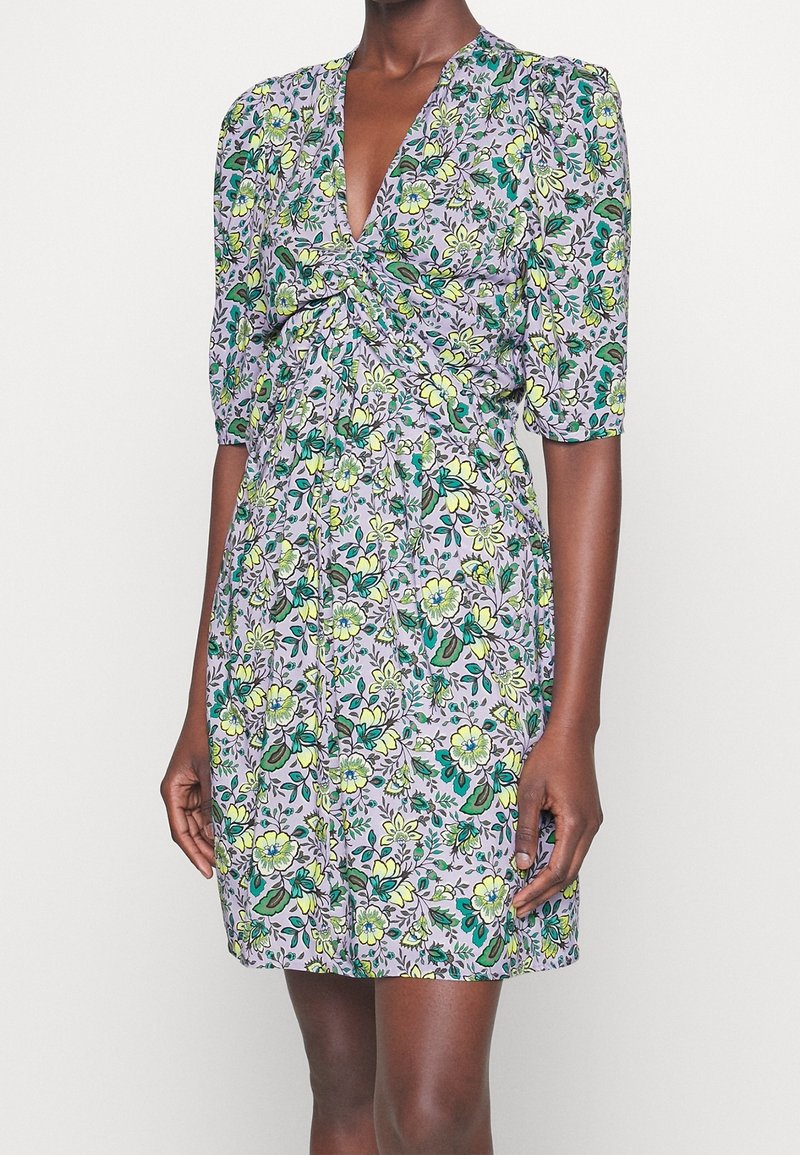 Femme portant une robe courte lilas avec un imprimé floral vert, jaune et blanc, avec un col en V et des manches bouffantes trois-quarts.
