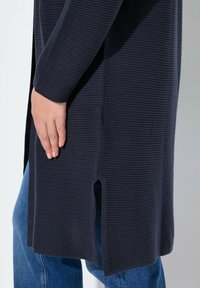 Cardigan en maille bleu marine foncé avec une surface texturée, des fentes latérales et des manches longues, associé à un jean en denim bleu.