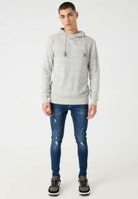 Sudadera con capucha gris claro con un patrón texturizado, cordones y detalles marrones. Combinada con vaqueros azules ajustados desgastados y zapatillas deportivas grises.