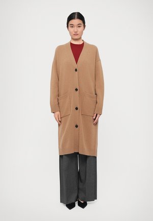 Cardigã longo, cor camelo, com decote em V, punhos canelados e dois bolsos frontais. Apresenta botões pretos e uma textura de malha suave. Combinado com calças cinzentas de perna larga e sapatos pretos.