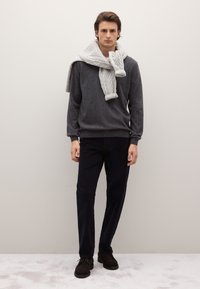 Grauer Strickpullover, hellgrauer Kabelstrickschal, dunkle Hose und braune Wildlederschuhe. Das Model steht vor einem schlichten Hintergrund.