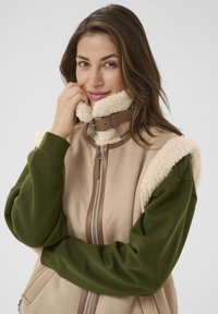 Beige fleece vest med grønne lange ærmer, der har en høj krave foret med lysebrunt kunstpels og en frontlynlås. Brune detaljer på hardware.