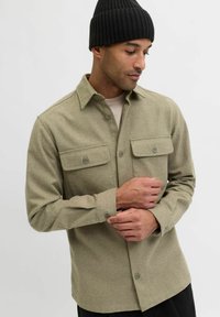 Olivengrønn skjorte med knappelukking og to brystlommer, laget av et teksturert stoff, med klassisk krage og knappede mansjetter. Tilbehøret inkluderer en svart strikket lue.