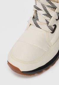 Cream-colored sneaker with a padded, smooth upper and black outsole. Features a textured cream and black striped lace for closure.