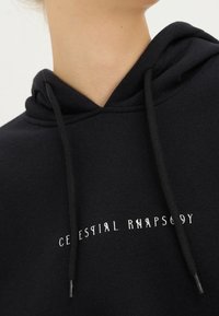 Sweat-shirt à capuche noir avec une texture douce, des cordons de serrage réglables, et le texte "CELESTIAL RHAPSODY" brodé en blanc sur la poitrine.