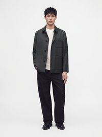 Homme portant une veste gris foncé boutonnée, une chemise blanche, un pantalon noir et des chaussures noires, debout devant un fond blanc uni.