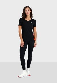 Black short-sleeve t-shirt with an orange side panel and small logo, paired with black leggings and white athletic shoes with red accents.