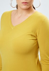 Gelbes V-Ausschnitt-Pullover aus weichem Strickstoff. Mit langen Ärmeln und Rippdetails am Ausschnitt und Saum. Glatte Textur.