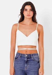 Top corto bianco a costine con scollatura a V e spalline sottili. Presenta un dettaglio annodato intorno alla vita. Abbinato a jeans blu a vita alta.