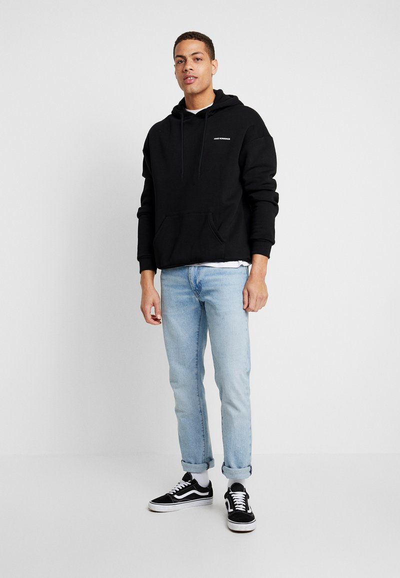 Czarna bluza z kapturem z przednią kieszenią, szare logo tekstowe, jasnoniebieskie dżinsy i czarne sneakersy z białymi podeszwami. Prosty, casualowy design.