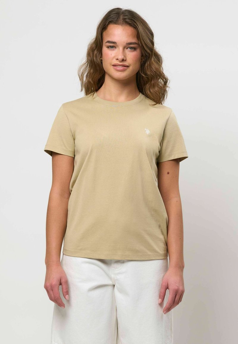 Jeune femme aux cheveux bruns ondulés portant un t-shirt beige à manches courtes et un pantalon blanc, debout devant un fond clair uni.