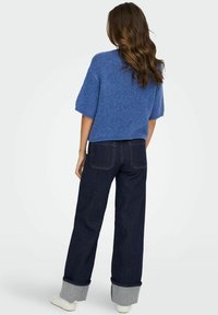 Zils adīts īsām piedurknēm džemperis ar brīvu piegriezumu, savienots ar tumšām džinsu biksēm ar platām kājām, kurām ir sarullēts apakšmalas vīlējums un kontrastējoša dūkošana.