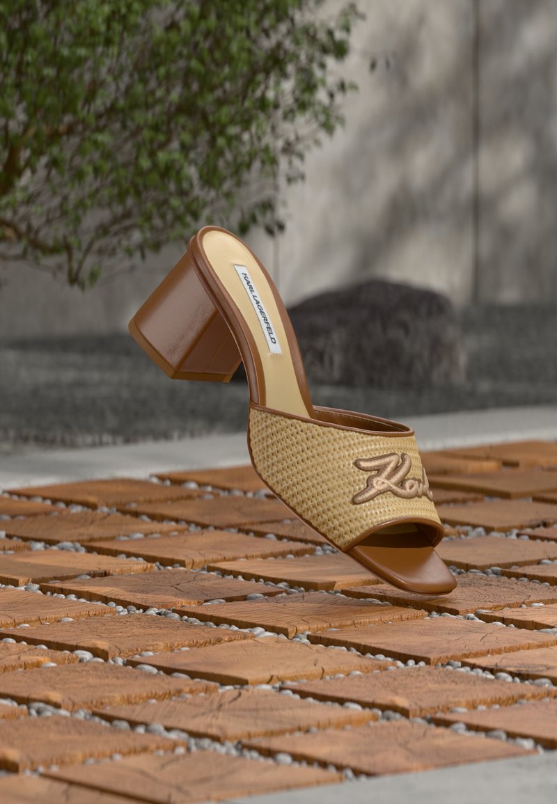 Καφέ mule σανδάλι με τακούνι, πλεκτό λουρί και κέντημα "Karl", τοποθετημένο πάνω σε ξύλινες πλάκες με φόντο πράσινο φυτό και πέτρινο τοίχο.