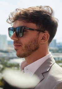 Černé a šedé sluneční brýle s lesklým povrchem a zrcadlovými čočkami, které nosí osoba v světle šedém obleku a růžové košili na pozadí města.