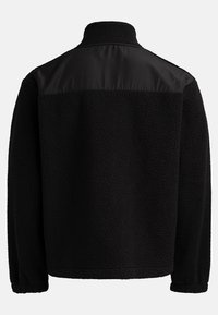 Veste en polaire noire avec un col haut et un panneau en tissu lisse sur le haut du dos, vue de l'arrière sur un fond uni.
