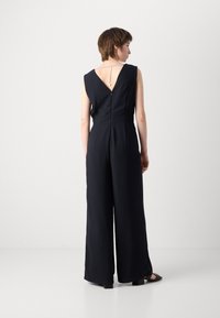 Mørkeblå jumpsuit med vide ben, V-formet ryggdesign og en sølvkjede detalj ved halslinjen. Glatt stoff med en skreddersydd passform.