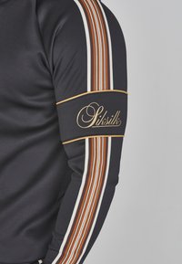 Top atlético negro con mangas a rayas en naranja, blanco y marrón y un diseño ajustado. Presenta un logo dorado en una banda para el brazo a juego.