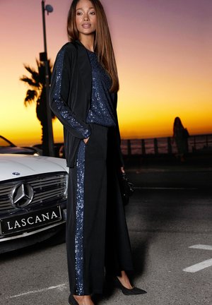 Outfit nero con accenti di paillettes. La parte superiore presenta paillettes blu, mentre i pantaloni a gamba larga hanno paillettes lungo il lato. Blazer nero e scarpe.