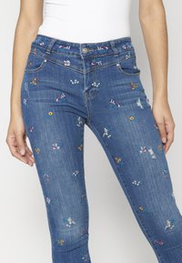 Vyšívané modré denimové džíny s květinovými vzory v různých barvách, slim fit, s mírně opraným detailem v oblasti stehen.