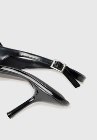 Svarte patenterte skinnhøye sandaler med spiss tå, ankelrem og sølvfarget spenn. Glatt tekstur og elegant design.