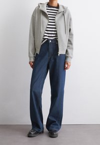 Un sweat à capuche zippé gris sur une chemise rayée noire et blanche, associé à un jean en denim foncé à taille haute et jambes larges, avec des chaussures noires.