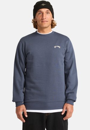 Marineblå sweatshirt i et blødt, struktureret stof med ribbede manchetter og kant, samt et lille hvidt logo på brystet. Parret med en sort beanie.