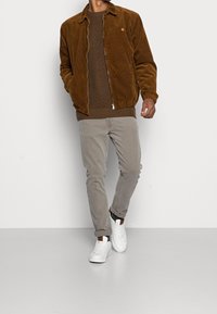 Brun kordjacka med dragkedja, ribbat brunt tröja, ljusgrå byxor och vita sneakers. Enkelt mönster med texturerad tyg.