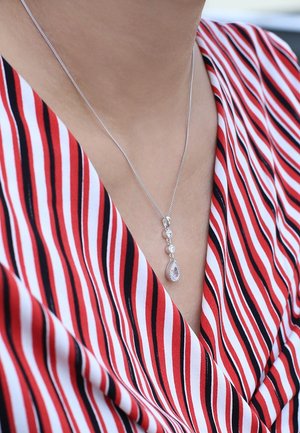 Zilveren ketting met een druppelvormige hanger met een edelsteen en drie ronde stenen, gedragen op een gestreepte rood-witte blouse.