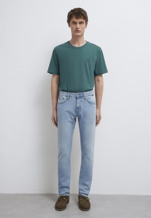 Ein Mann steht vor einer weißen Wand, trägt ein türkises Kurzarmhemd, hellblaue Jeans und braune Schnürschuhe und schaut mit den Armen an den Seiten nach vorne.