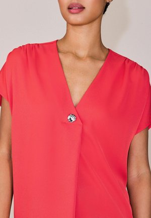 Femeie purtând o bluză cu mâneci scurte de culoare coral strălucitor, cu decolteu adânc în V și un singur nasture decorativ argintiu în față.