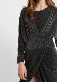 Czarna sukienka z długimi rękawami, marszczonym paskiem w talii i drapowaną spódnicą. Posiada metaliczne srebrne paski oraz boczny rozcięcie, co tworzy teksturowany wygląd.