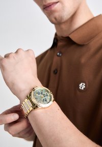 Ceas cronograf din aur cu un cadran albastru și accente de diamante, având o brățară din metal groasă. Purtați pe un încheietură, asortat cu un tricou polo maro.