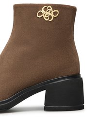 Stivale alla caviglia in suede marrone con punta rotonda, tacco a blocco nero e elemento decorativo dorato sul lato. Texture morbida e design pulito.