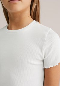 Biała koszulka z krótkim rękawem, prążkowana, z okrągłym dekoltem i subtelnie falbaniastymi krawędziami rękawów, wykonana z miękkiego, teksturowanego materiału.