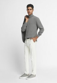 Szary sweter z golfem, kremowe spodnie i szare sneakersy. Model trzyma czarną marynarkę na lewym ramieniu. Tło jest jednolicie białe.