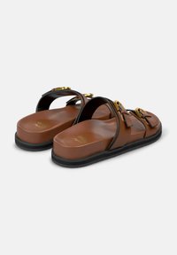 Sandalias de piel marrón tipo slip-on con acentos negros, detalles de hebilla y una suela plana negra, con una textura lisa y brillante.